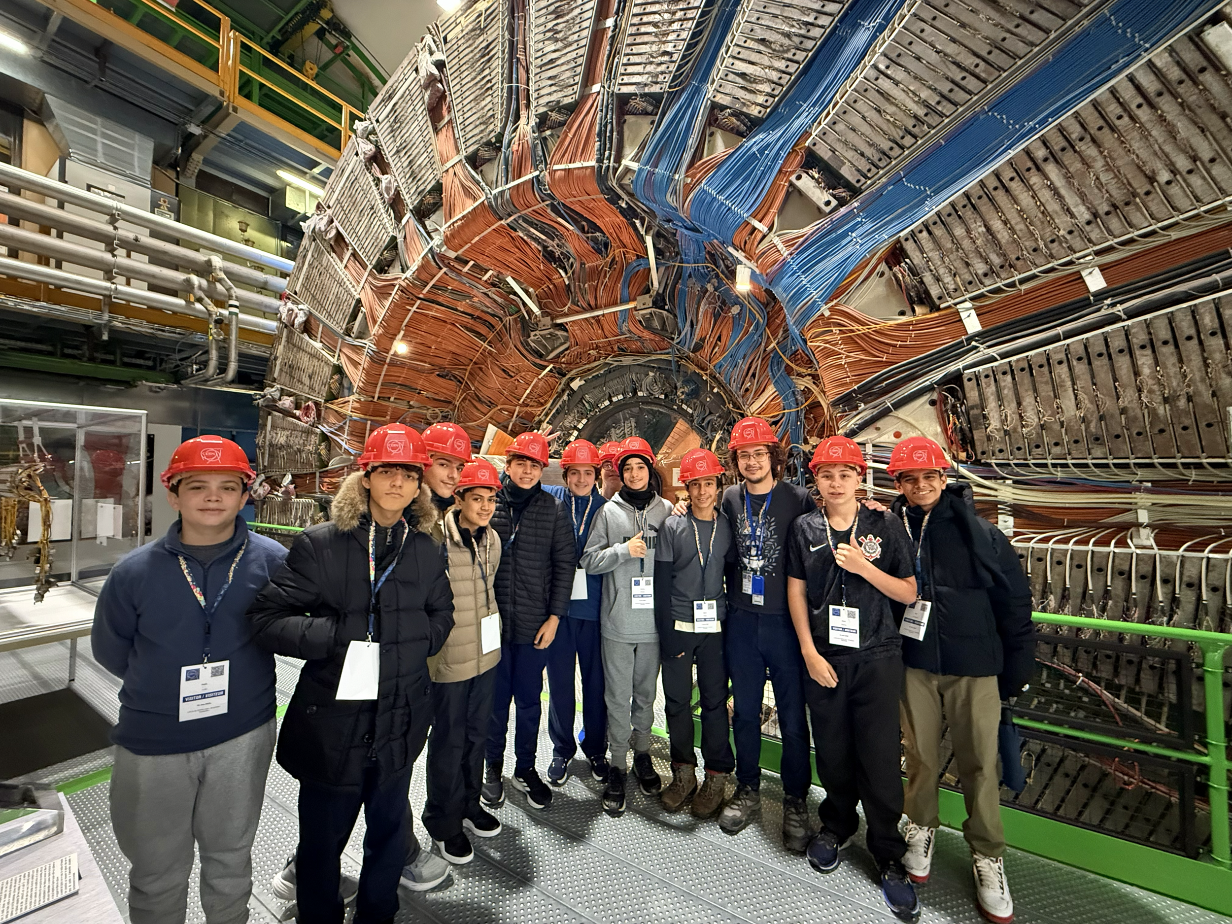 Descrição da imagem: foto de um grupo de estudantes à frente de uma enorme estrutura circular repleta de cabos.