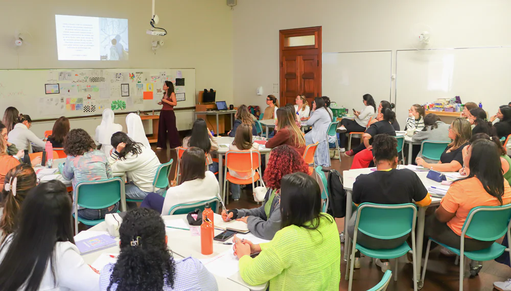 Descrição da imagem: foto de uma sala de aula repleta de educadoras. Uma delas está à frente do grupo, perto de uma lousa.