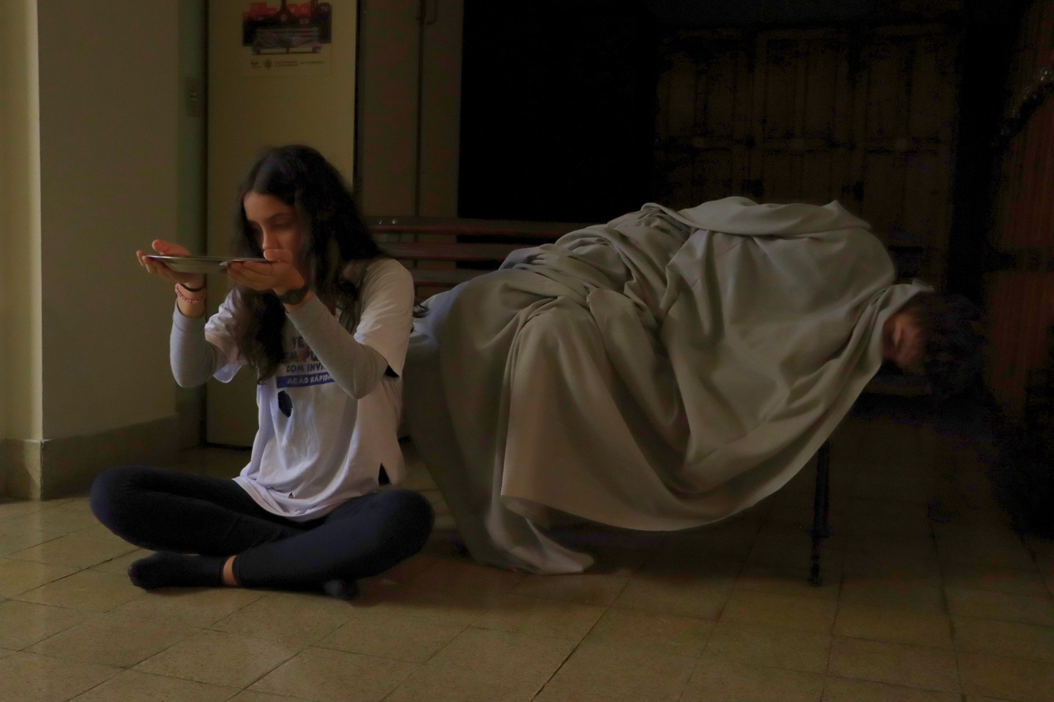 Descrição da imagem: foto de uma estudante sentada no chão, segurando um prato vazio, e um aluno deitado num banco, coberto com cobertor.