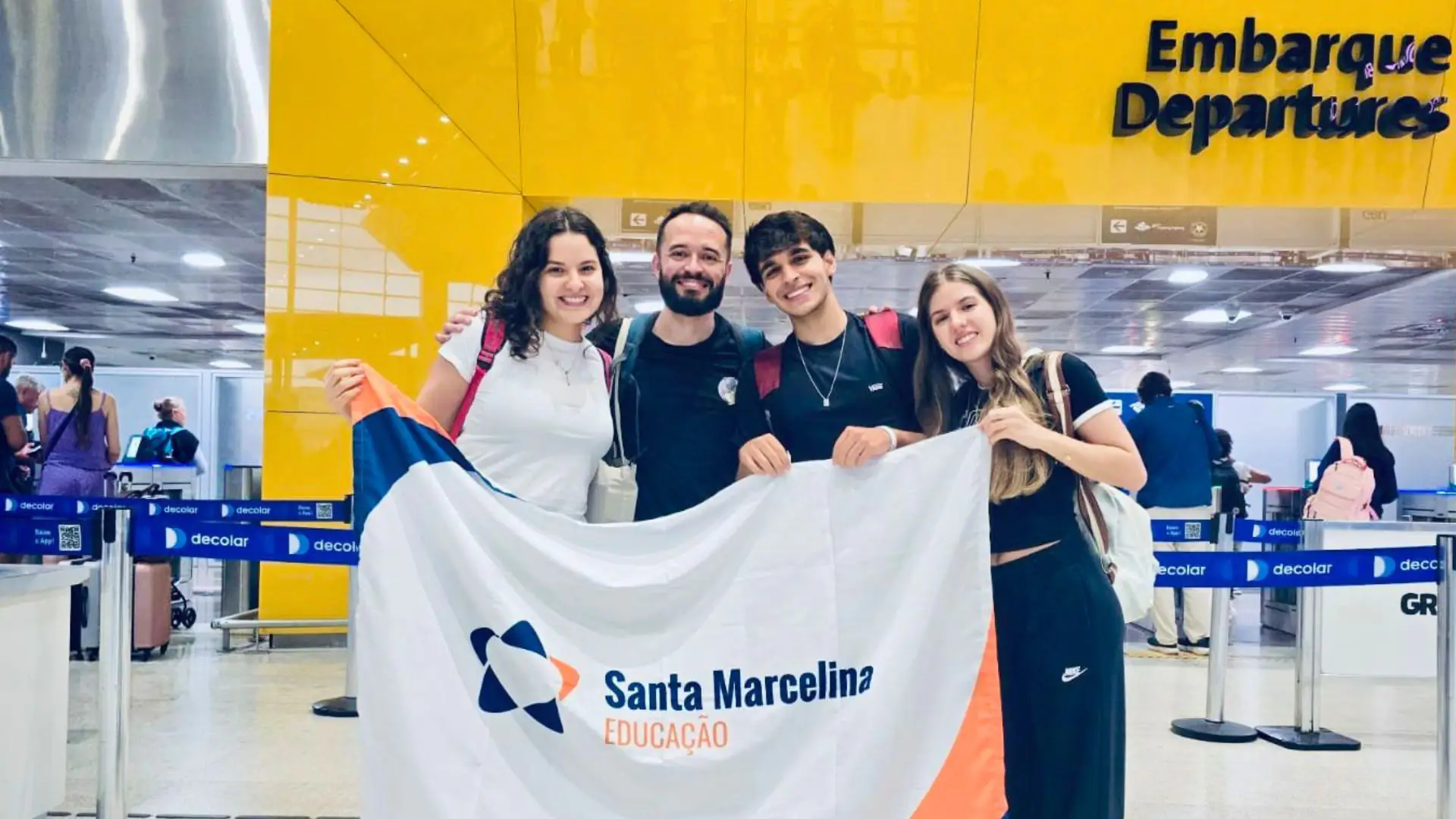 Descrição da imagem: foto de quatro pessoas segurando uma bandeira da Rede Santa Marcelina em frente a um portão de embarque.