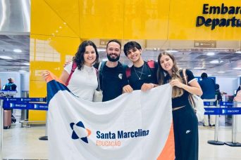 Descrição da imagem: foto de quatro pessoas segurando uma bandeira da Rede Santa Marcelina em frente a um portão de embarque.