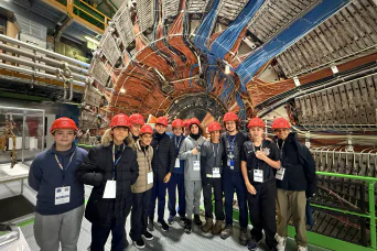 Descrição da imagem: foto de um grupo de estudantes à frente de uma enorme estrutura circular repleta de cabos.